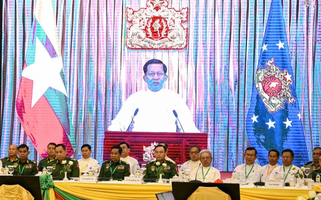 Myanmar officials attending a meeting with a screen showing a leader.
