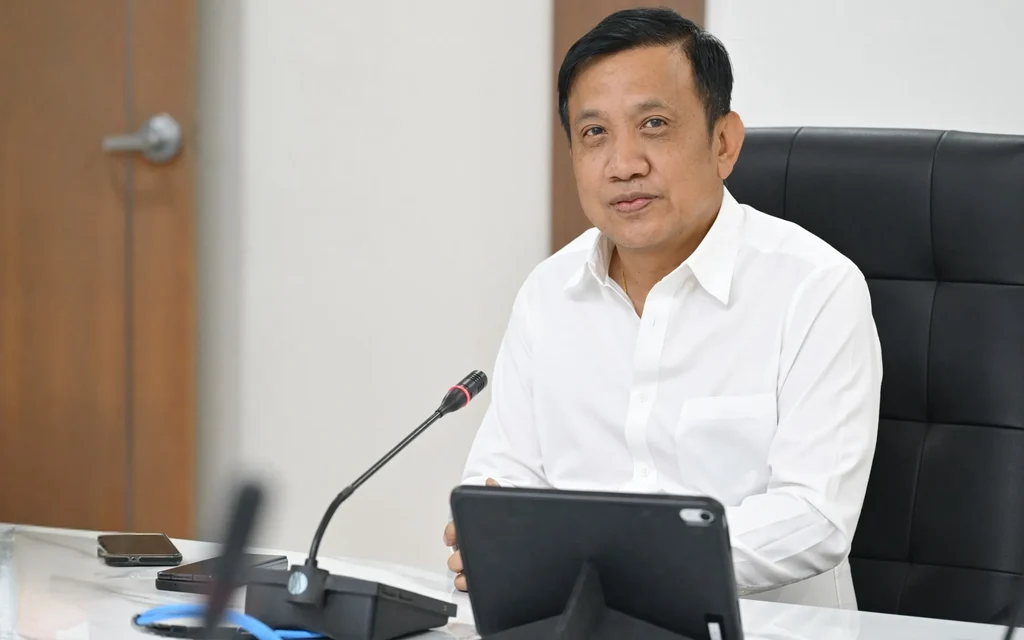 A man in a white shirt seated at a conference table with a tablet and microphone.