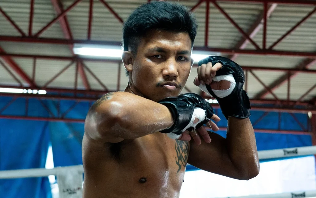 A shirtless Muay Thai fighter poses with wrapped hands in a training ring.