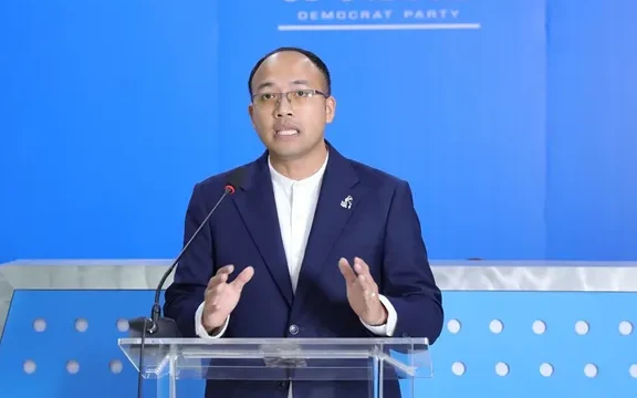 A Democrat Party representative speaks at a lectern against a blue backdrop.