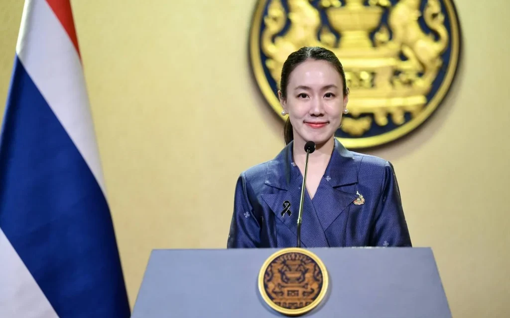 Woman speaks at a podium beside the Thai flag and official emblem.