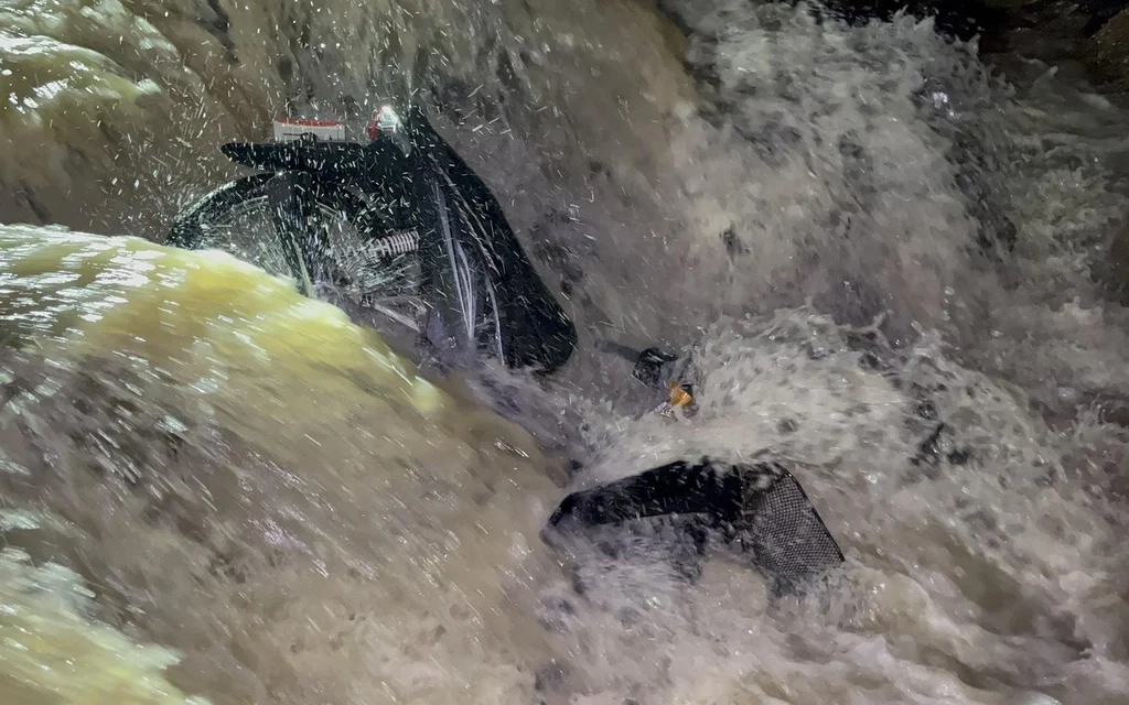 A motorcycle submerged in a fast-flowing flooded drainage channel.
