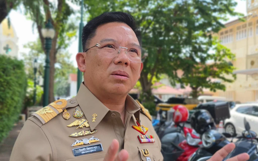 A uniformed Thai official speaks outside near parked motorcycles.