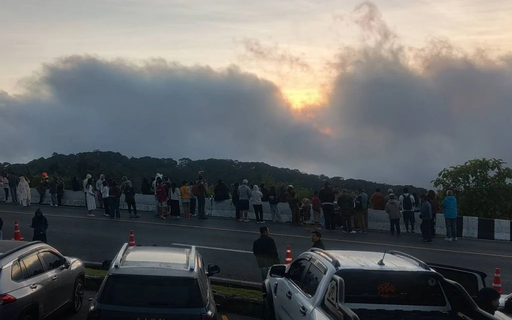 Tourists gather beside parked cars to watch a cloudy sunset over hills.