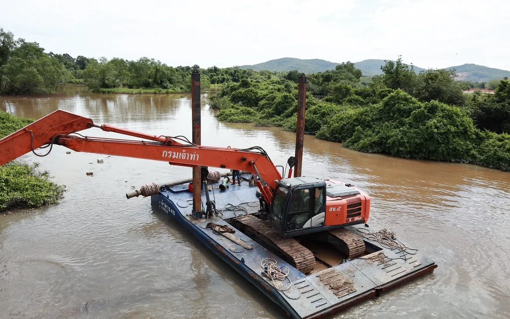 An orange excavator sits on a barge in a river surrounded by greenery.