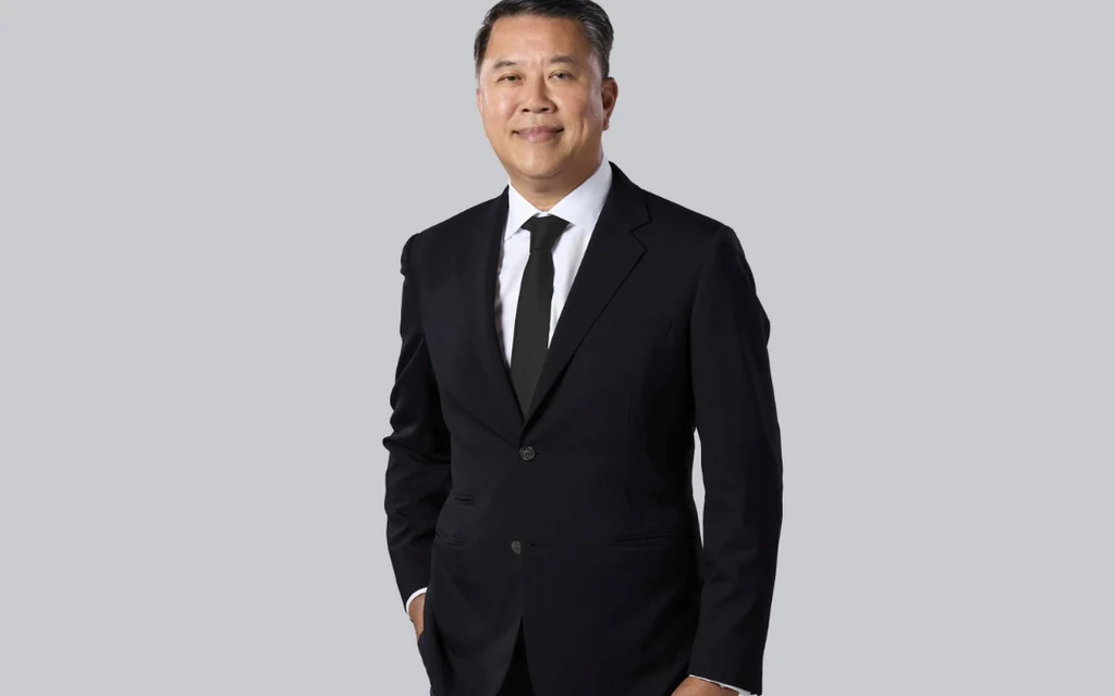 A man in a black suit stands against a plain gray background.