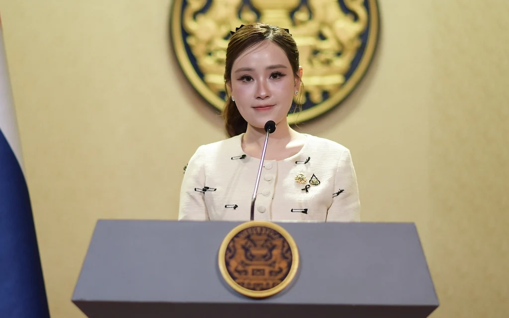 A government spokesperson speaking at a podium with the Thai flag.