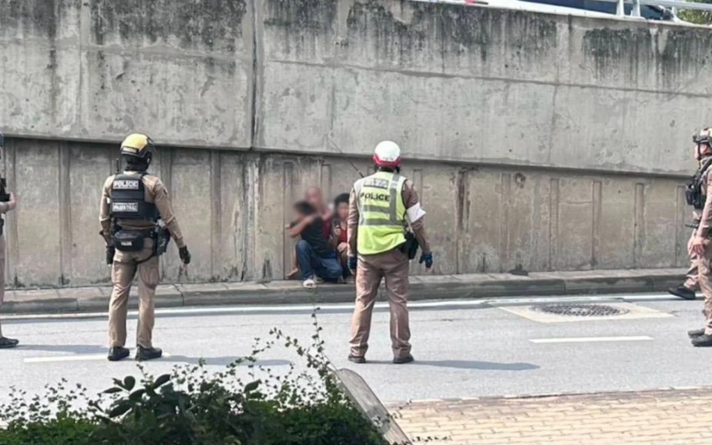 Police officers surround a blurred man and child beside a Bangkok wall.