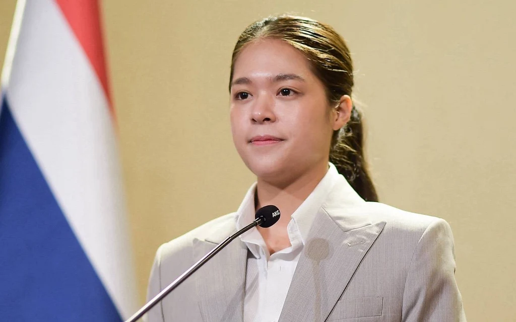 A woman speaks at a microphone beside a Thai flag.