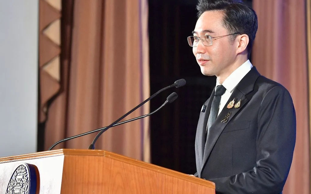 A man in a suit speaks at a wooden lectern in a formal hall.