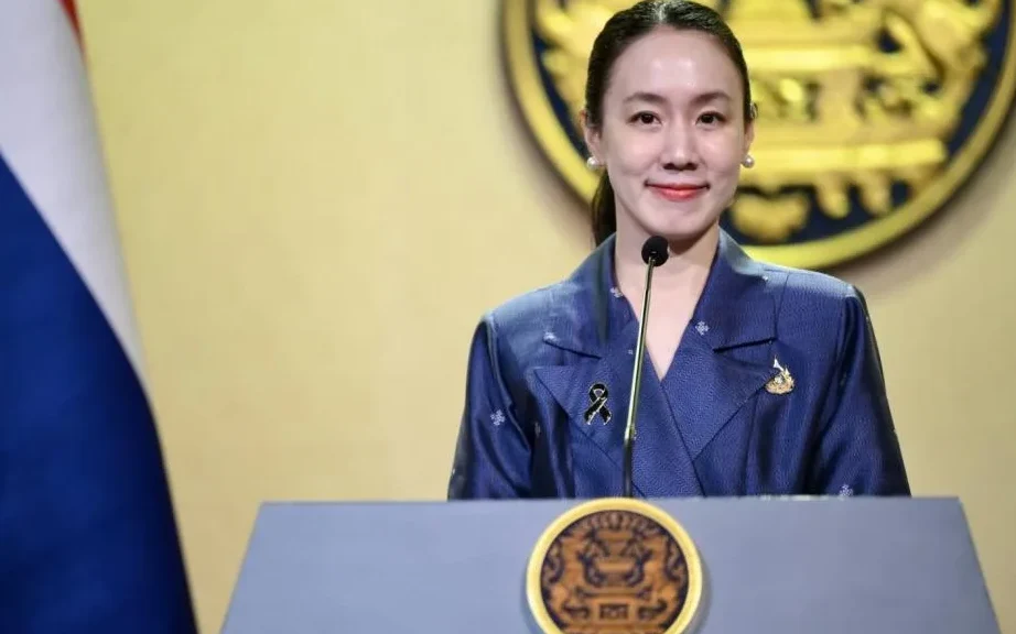 A woman speaking at a podium with a Thai government emblem.