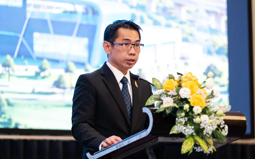 Man in a suit speaks at a podium beside flowers.