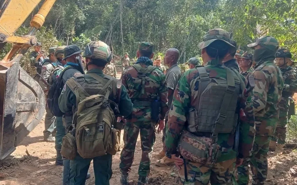 Soldiers gather beside an excavator on a wooded border road.