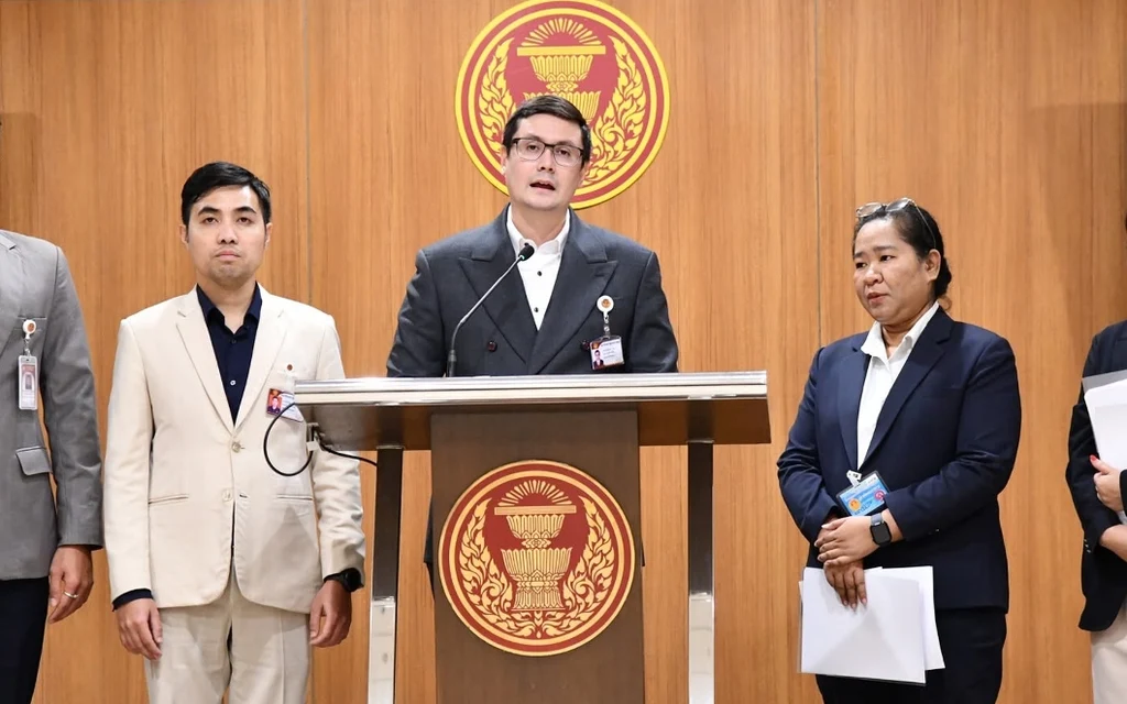 Officials speak at a podium in a wood-paneled assembly hall.