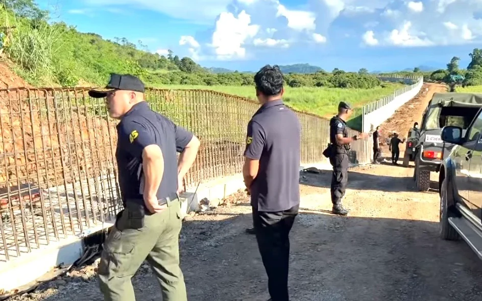 Officials inspect a border fence construction site beside parked vehicles.