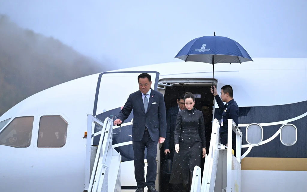Officials and a woman descend airplane stairs beneath an umbrella.