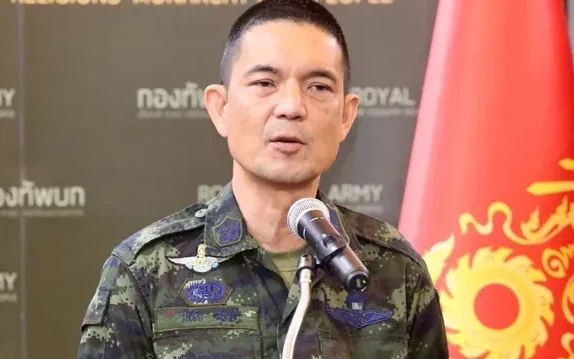 Thai military officer speaking at a microphone beside a military flag