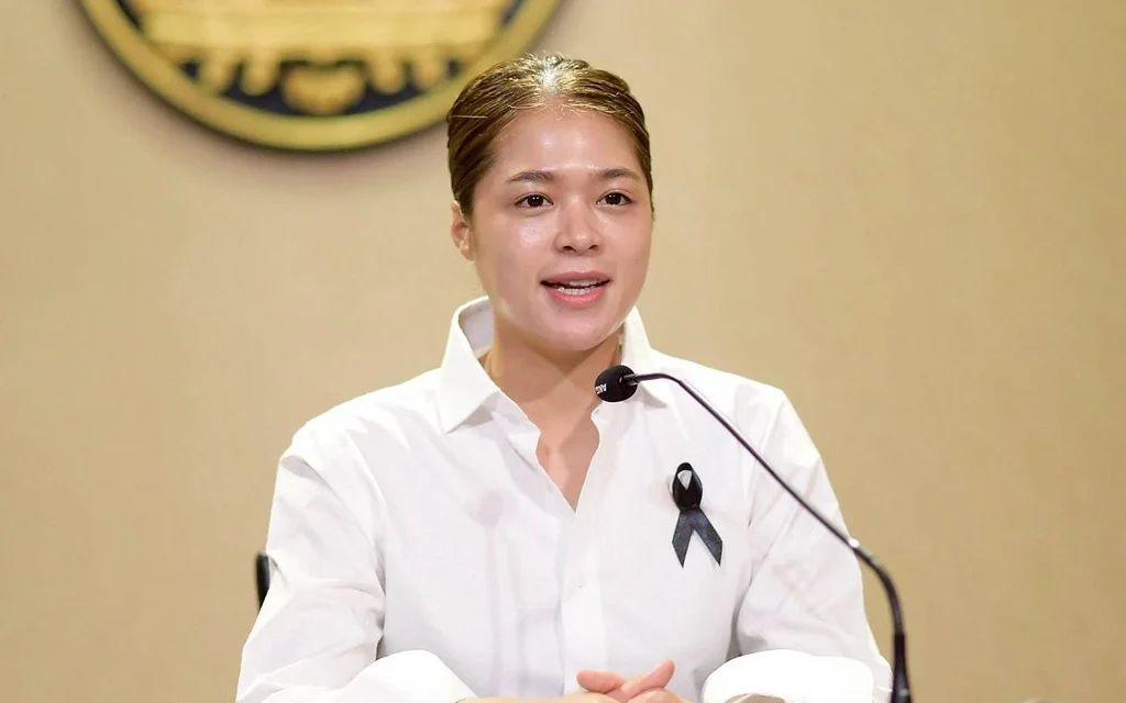 Woman speaking at a microphone, wearing a white shirt and black ribbon.