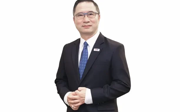 A man in a dark suit stands against a plain white background.