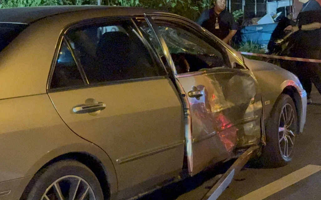 Damaged sedan at a nighttime motorcycle crash scene with bystanders nearby