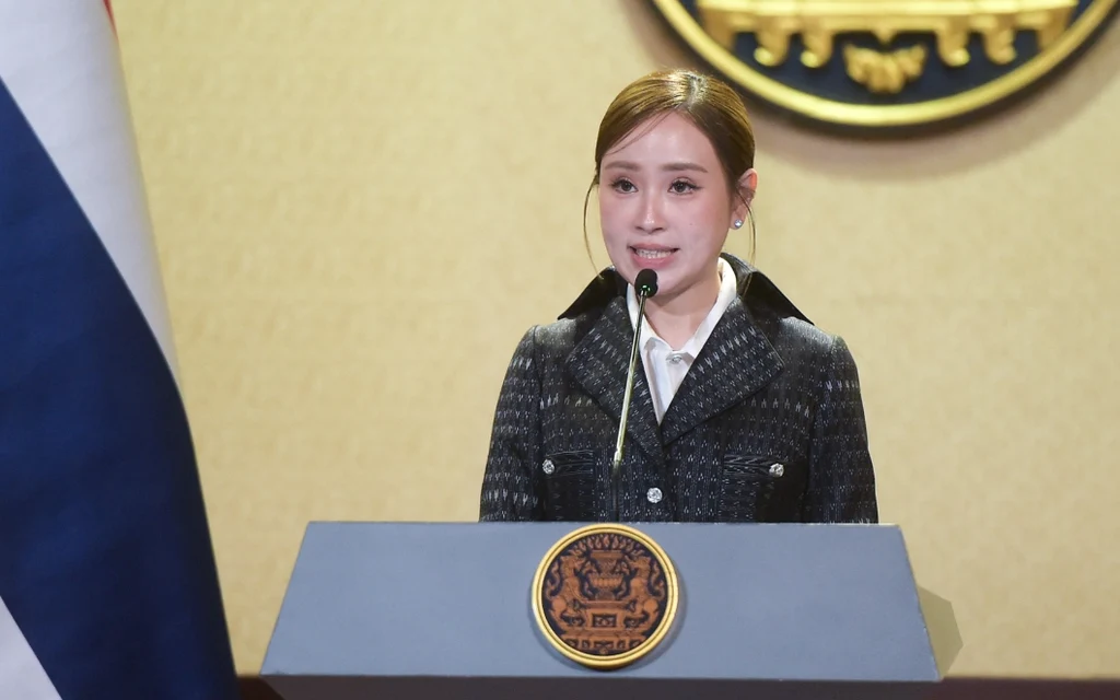 A woman speaks at a podium beside a Thai flag.
