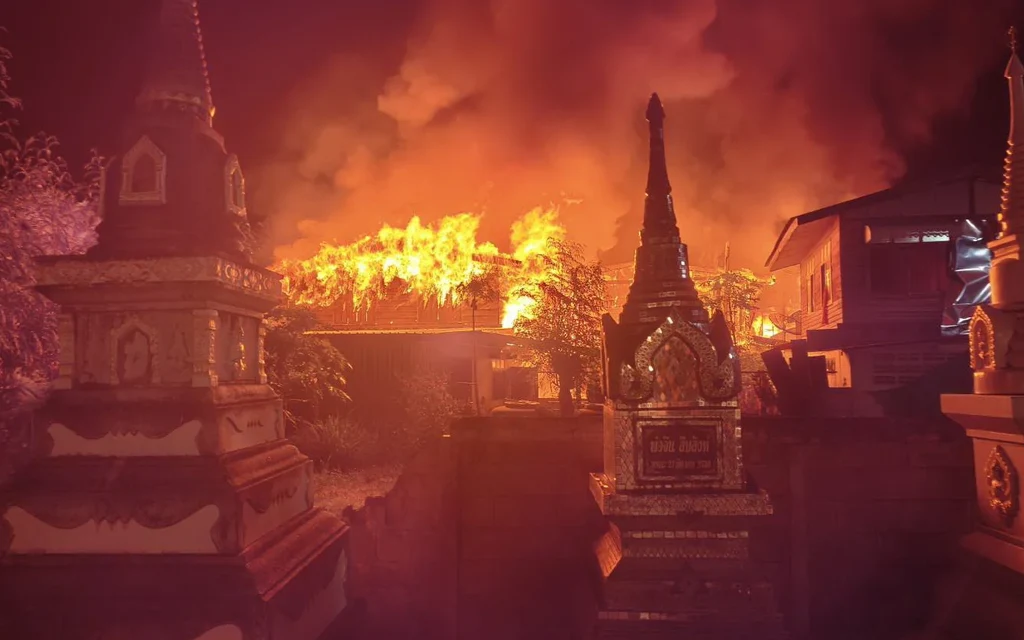 Fire burns houses behind Buddhist stupas in a nighttime community scene.
