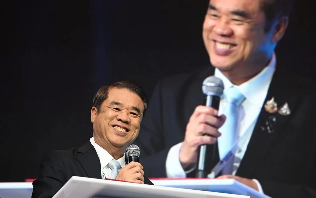A smiling man speaks at a lectern before a projected image of himself.