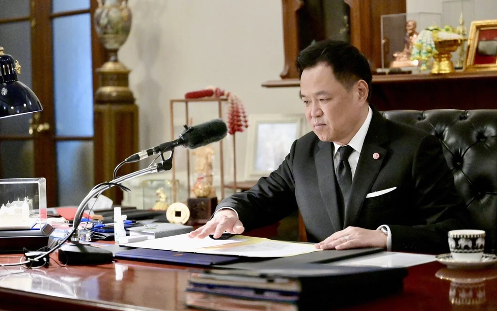A man in a suit speaks at a desk in an office.