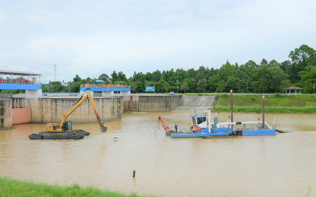 Excavator and workboat operate in muddy water near a flood-control structure.