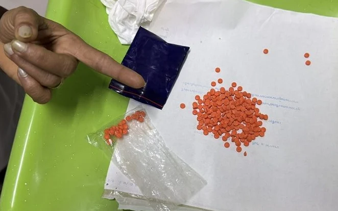 Orange yaba pills, plastic wrapping, and a pointing hand on a table.