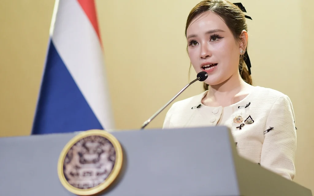 A government spokesperson speaking at a podium with a Thai flag.