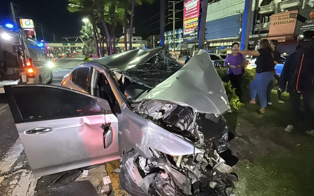 Severely damaged car at a nighttime crash scene with bystanders and emergency vehicles.