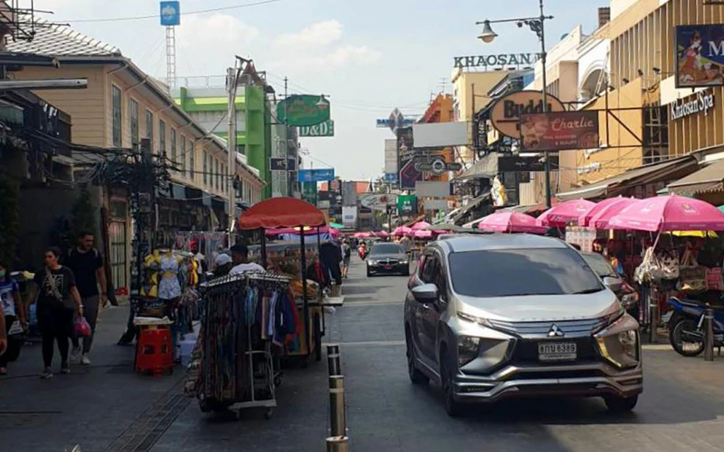 Street scene on Khaosan Road with shops, pedestrians, market stalls, and a car.