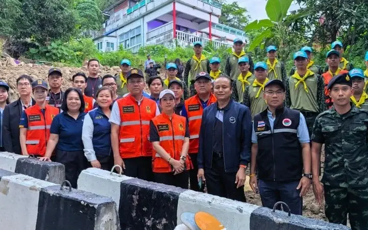 Officials and volunteers pose by flood barriers near a hillside building.