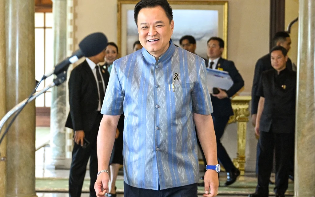 Thai politician Anutin walking through an ornate interior as reporters and staff stand behind him.