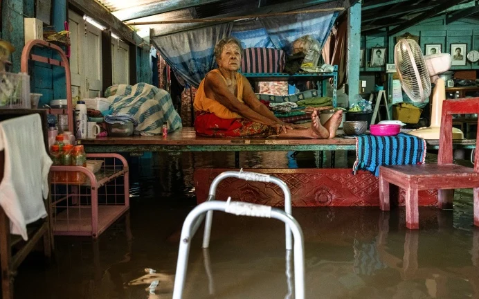 An elderly woman sits on a raised platform inside a flooded home.