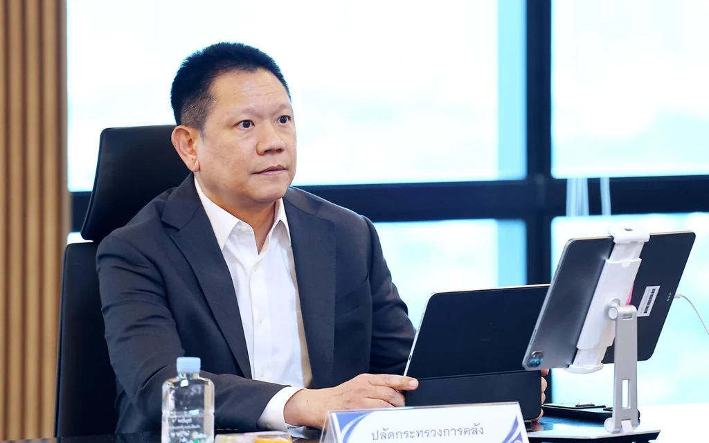 A suited official sits at a conference table with a tablet and nameplate.
