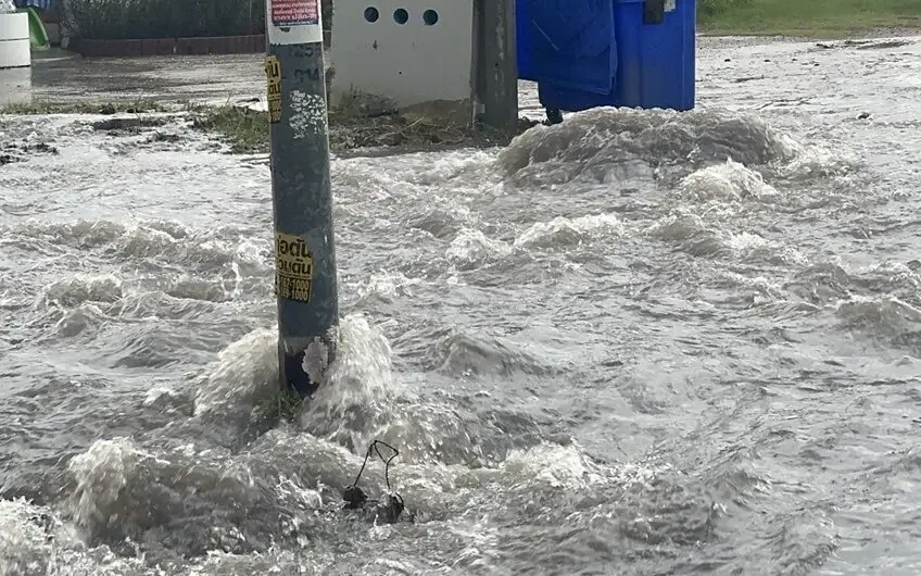 Floodwater rushes across a road beside a utility pole and bins.
