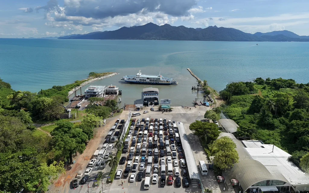 Aerial view of a ferry terminal with vehicles, a ferry, and surrounding sea.