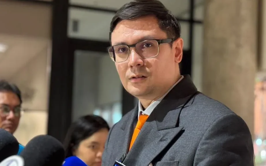 Man in a suit speaks to reporters surrounded by microphones.