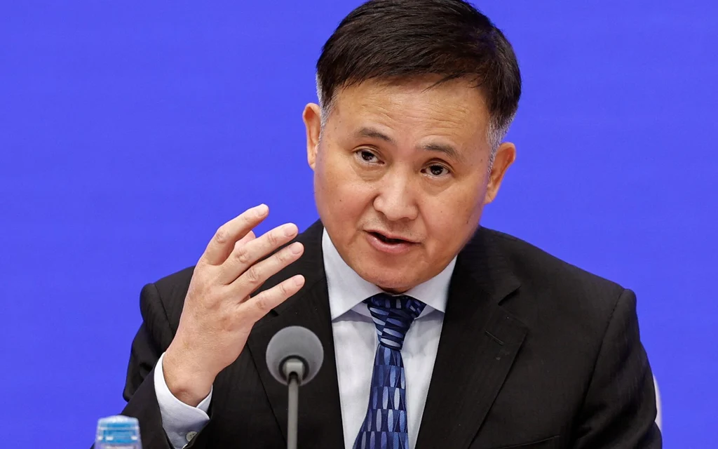 A man in a suit speaks at a press conference against a blue backdrop.