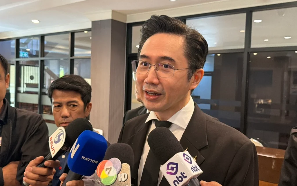 A suited official speaks to reporters and microphones in an indoor hallway.