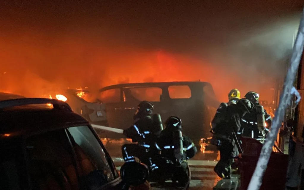 Firefighters battle a vehicle fire at a repair shop at night.