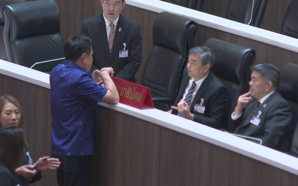 Politician greets seated officials inside the parliamentary chamber.