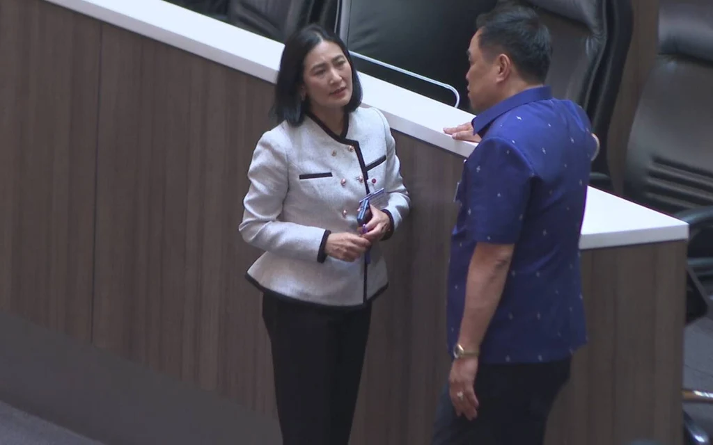 Two politicians talk beside seats in a parliamentary chamber.