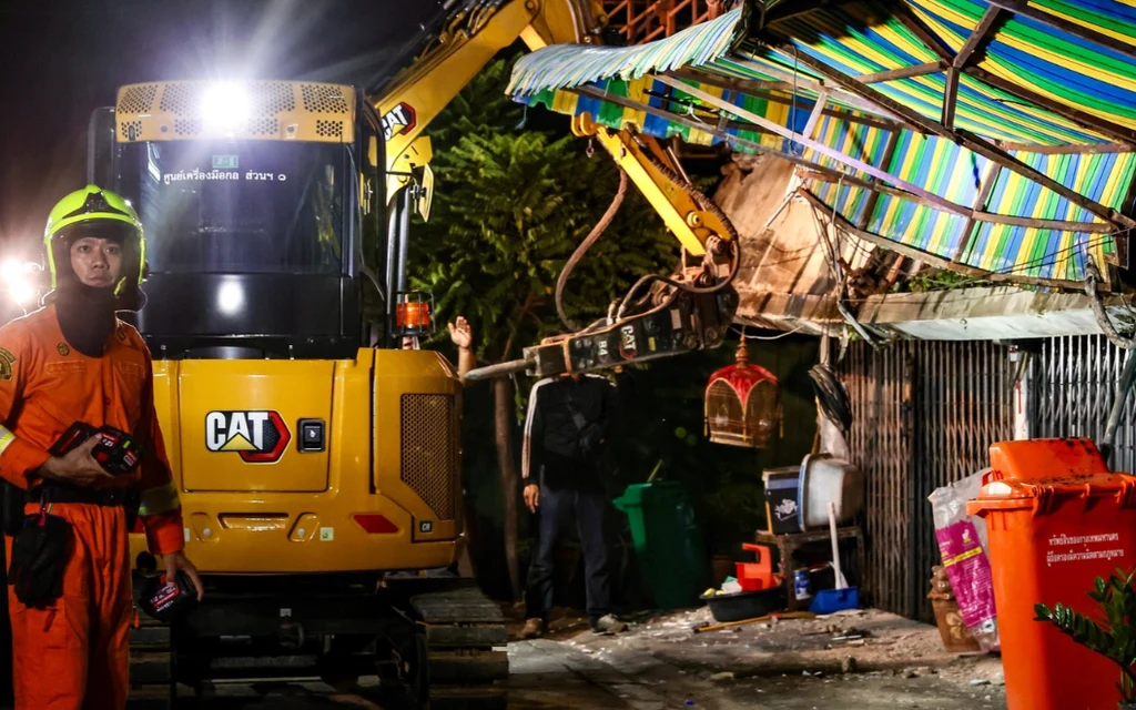 Emergency workers inspect a collapsed shophouse awning beside an excavator at night.