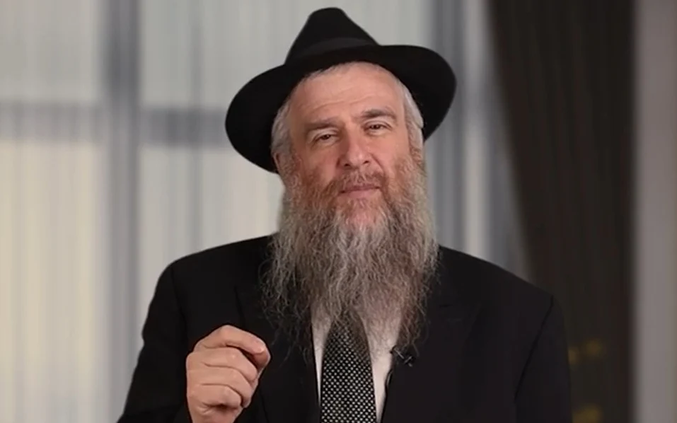 Bearded man wearing a black hat and suit, gesturing toward the camera.