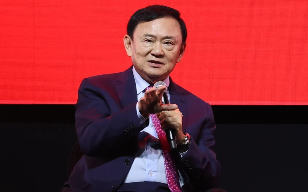 Thaksin Shinawatra speaking at an event with a red background.