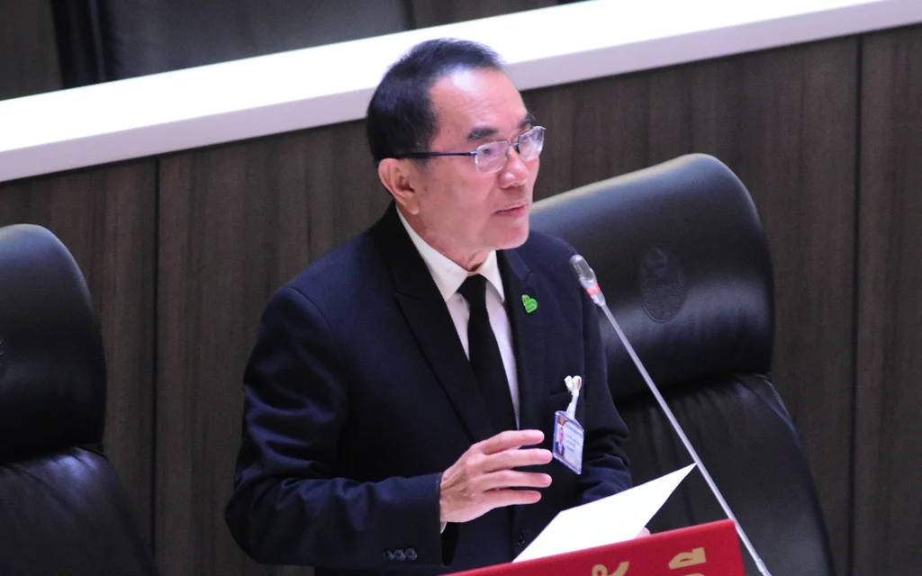 A suited man speaks at a parliamentary lectern beside a microphone.