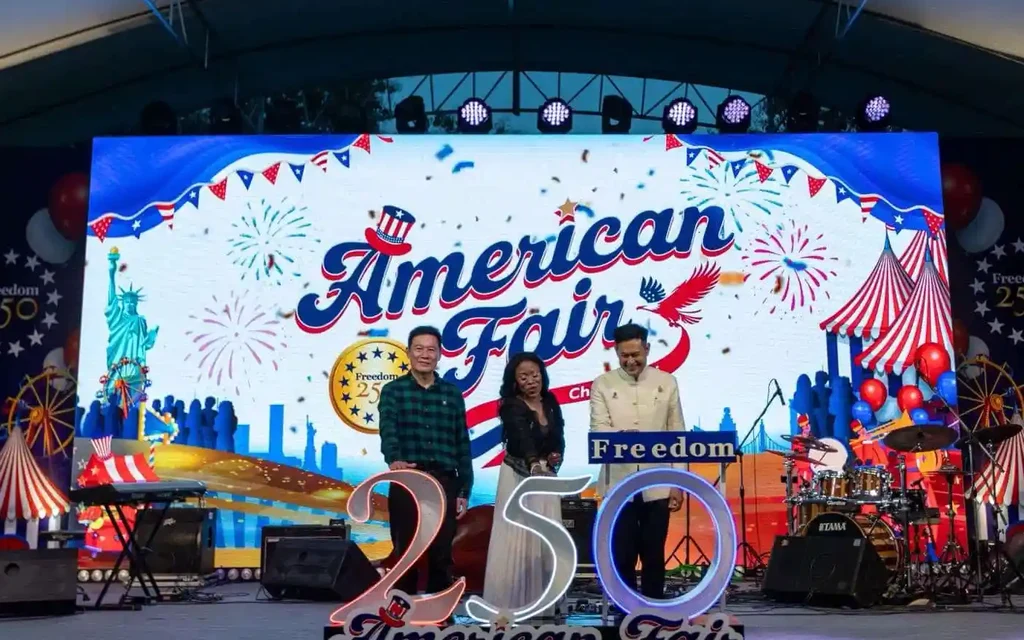 Three people pose onstage at an American fair anniversary celebration.